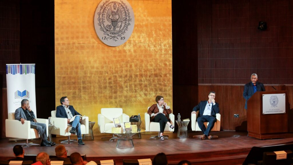 Image of the stage inside the auditorium with 4 panelists and Dr. Abdelwahhab El-Affendi speaking on thepodium