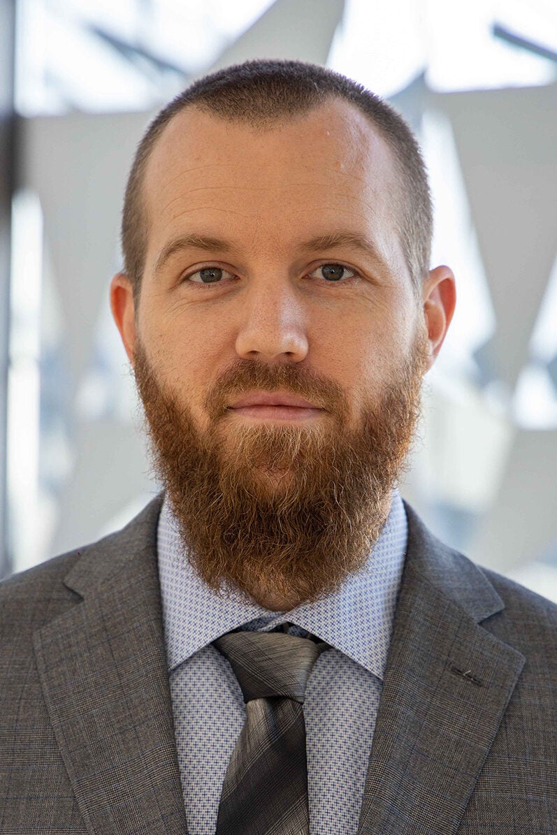 A man with a neatly trimmed beard and short hair, wearing a gray suit jacket, a light blue dress shirt, and a striped tie. He is looking directly at the camera with a neutral expression, with a blurred, abstract background behind him.