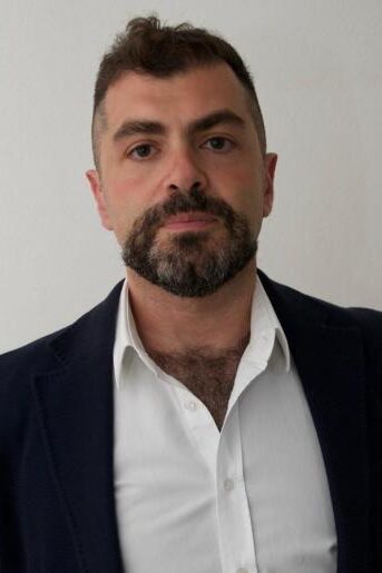 A man with short dark hair and a trimmed beard, wearing a dark blazer over a white collared shirt, standing against a plain light-colored background, looking directly at the camera with a neutral expression.