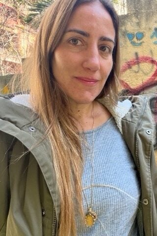 A woman with long, light brown hair, wearing a light green jacket over a gray top, standing outdoors. She is on a staircase with a graffiti-covered wall in the background, looking at the camera with a slight smile.